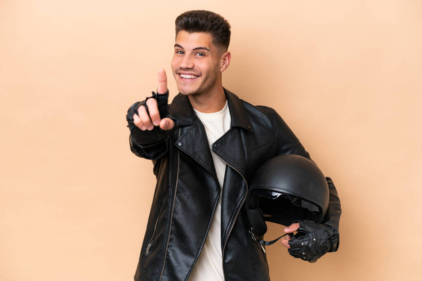 Jovem caucasiano com um capacete de motocicleta isolado no fundo bege mostrando e levantando um dedo - Foto, Imagem
