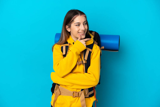 Молода альпіністка з великим рюкзаком ізольована на синьому фоні, дивлячись в сторону і посміхаючись
 - Фото, зображення