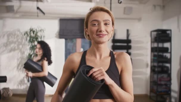Portré kaukázusi női edzőről, aki vidáman mosolyog a kamera előtt, miközben fitneszszőnyeget tart. A háttérben látható boldog nők aktívan beszélgetnek egymással kemény edzés után. - Felvétel, videó