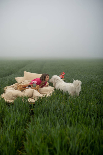 Jovem mulher gosta de tempo da manhã, deitada na cama com seu cachorro bonito e tomando café da manhã doce no campo verde ao ar livre - Foto, Imagem