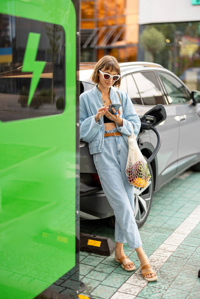 Mujer joven se para con un teléfono inteligente y una bolsa de malla con algo de comida, esperando a que su coche eléctrico se cargue en una estación pública al aire libre. Concepto de estilo de vida sostenible y energía verde - Foto, Imagen