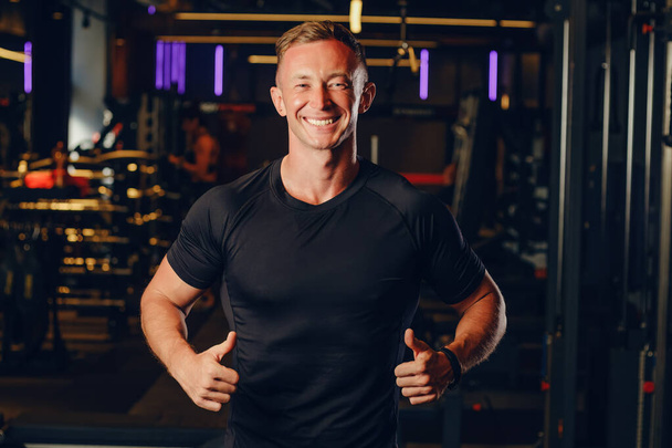 The guy athlete in a black T-shirt without inscriptions shows a thumbs up gesture and looks at the camera smiling. Place for text or design, mockup, copy space. - Photo, Image