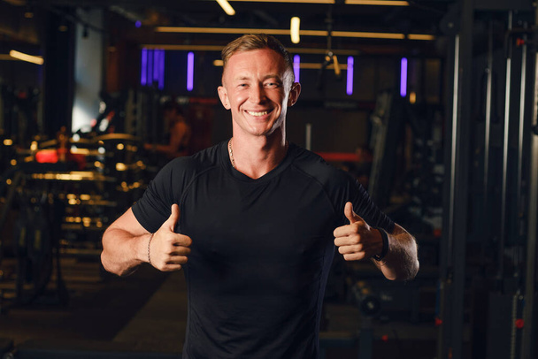 The guy athlete in a black T-shirt without inscriptions shows a thumbs up gesture and looks at the camera smiling. Place for text or design, mockup, copy space. - Photo, Image