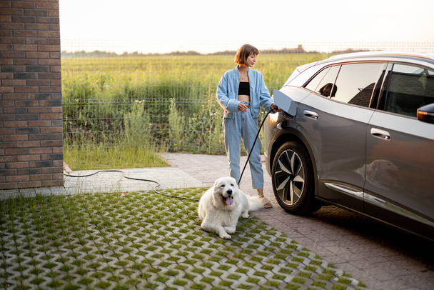 Mulher carrega carro elétrico, de pé com seu cão branco bonito perto de sua casa no pôr do sol. Conceito de energia verde, sustentabilidade e estilo de vida moderno - Foto, Imagem