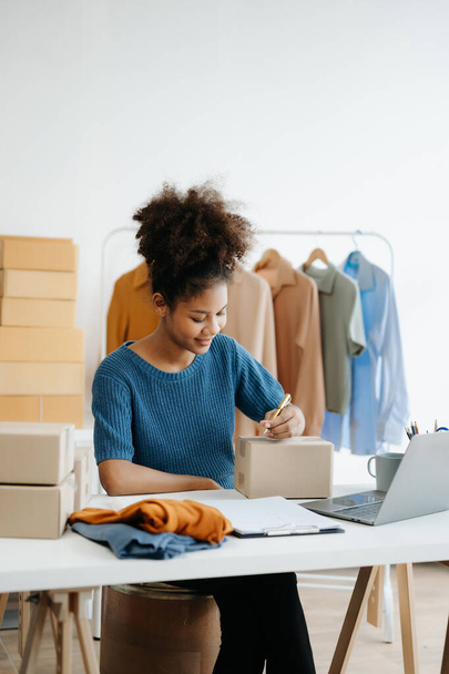 Startup small business SME. Entrepreneur African woman receiving and checking online orders. Young female small business owner packing parcels to ship them to customers - Zdjęcie, obraz