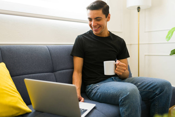 Felice giovane uomo che lavora da casa utilizzando il computer portatile mentre indossa una maglietta finta utilizzando una tazza di caffè - Foto, immagini