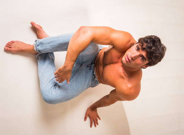 Young handsome shirtless man sitting on floor in studio against white, looking up - Foto, immagini