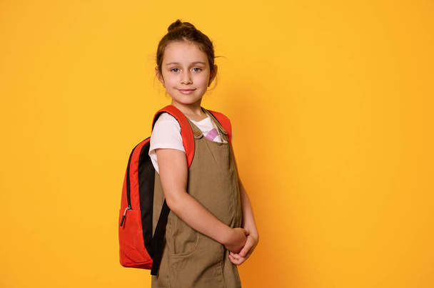Retrato seguro de una adorable niña caucásica de 6 años, niño de escuela primaria, estudiante de primer grado con mochila, sonriendo tiernamente mirando a la cámara, aislado en el fondo del estudio de color amarillo - Foto, imagen