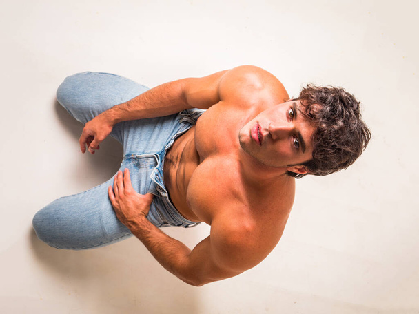 Young handsome shirtless man sitting on floor in studio against white, looking up - Foto, afbeelding
