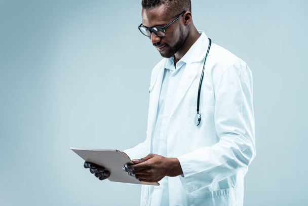 fókuszált modern orvos, fekete férfi ápolt szakállal, digitális technológiát használ a jobb betegellátás érdekében. Tartja tabletta, visel fehér kabátot, több fokozatú, jóképű - Fotó, kép
