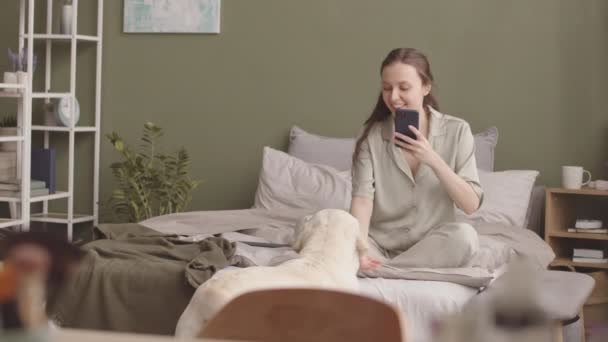 女の子は居心地の良い朝に自宅でベッドに座っている間,かわいいラブラドールのリトリーバーのスマートフォンで写真を撮ります - 映像、動画