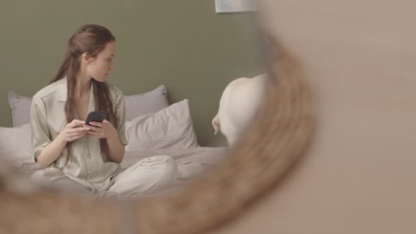 Lány kényelmes pizsamában sms-ezés okostelefonon, miközben pihen az ágyban a kutyájával a lusta reggelen - Felvétel, videó