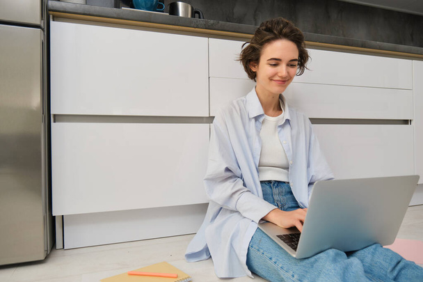 Молодая IT-девушка, женщина работает внештатно из дома, сидит на полу с ноутбуком. Девочка-студентка делает домашнее задание онлайн, пишет на клавиатуре компьютера. - Фото, изображение