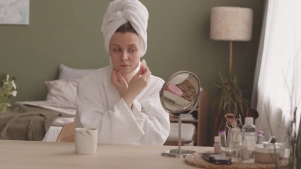 Joven atractiva mujer caucásica en albornoz blanco y toalla en su cabeza haciendo masaje facial con gua sha de cuarzo en el dormitorio - Metraje, vídeo