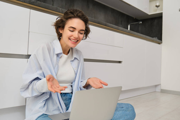 Portrait de jeune fille freelance se connecte à une réunion en ligne, chats vidéo sur ordinateur portable, se trouve sur le sol de la cuisine et aime sa conversation. - Photo, image