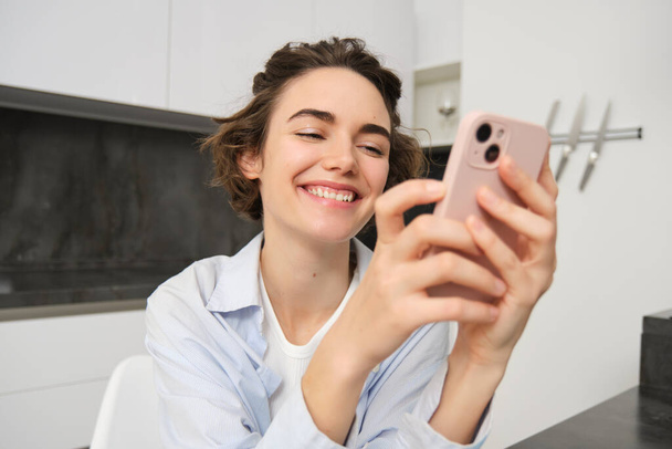 Portrét brunetky žena chatuje na mobilním telefonu, používá aplikaci smartphone z domova, objednávky doručení, zatímco sedí v kuchyni. - Fotografie, Obrázek