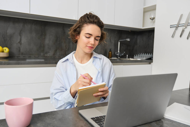 Femme qui travaille prend des notes, écrit des informations sur le carnet, assis avec un ordinateur portable à la maison. - Photo, image
