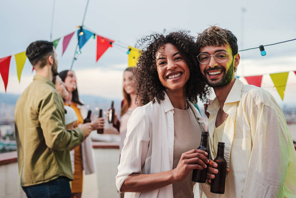 Portrait of young adult multiracial couple smiling, laughing and looking at camera holding beer bottles. Two people staring front on a party celebration with their best friends talking at background - Photo, Image
