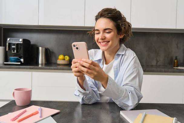 Teknoloji ve yaşam tarzı. Genç bir kadın evinde oturuyor, mutfağında akıllı telefon kullanıyor ve gülümsüyor.. - Fotoğraf, Görsel
