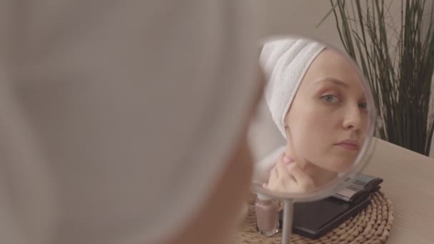 Joven hermosa mujer caucásica en albornoz blanco y toalla en su cabeza aplicando crema en su cuello mientras hace rutina de cuidado de la piel delante del espejo en el dormitorio - Metraje, vídeo