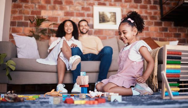 Happy, playing and a child with building blocks and parents for relax, education and learning in a house. Smile, playful and a girl kid with toys and a mother and father in a living room as family. - Photo, Image