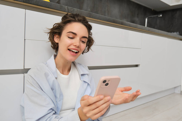 Portrait of happy young woman, shows her brand new kitchen, sits on floor, points at furniture and smiles, video chats on smartphone. Technology and lifestyle concept - Photo, Image
