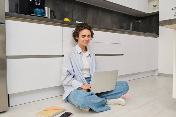 Mujer joven estudiando en el suelo. Chica con portátil trabaja desde casa, tipos en el ordenador, sonríe y se siente creativo. - Foto, Imagen