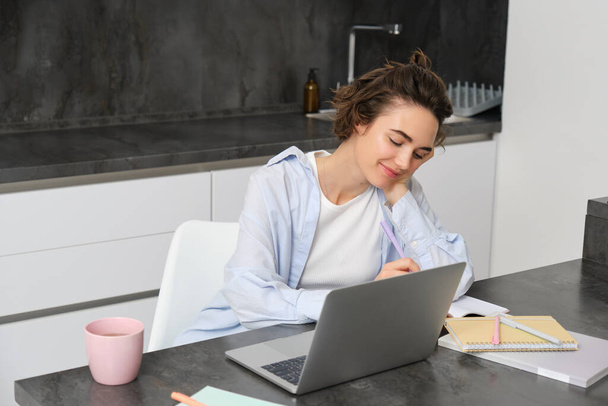 Image of young productive woman works from home, does distance learning, learns online course on her laptop in kitchen, writes down information, watches webinar, attends work meeting remotely. - Photo, Image
