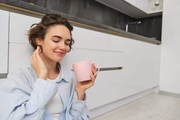 Kényelem és otthon koncepció. Fiatal nő élvezi a reggeli csésze kávét, iszik friss teát és mosolyog a kényelem és a kikapcsolódás, ül a konyha padlóján rózsaszín bögre. - Fotó, kép