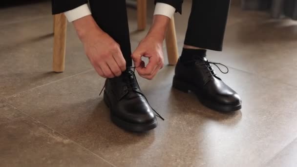 Een man in een pak bindt schoenveters op zwarte leren schoenen op een houten parket achtergrond zakelijke stijl. voorbereiding van een bruiloft of zakelijke bijeenkomst. middelen voor de verzorging van leren schoenen - Video