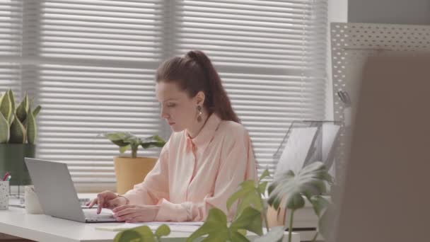 Молода кавказька жінка в довгому високому хвіст сидить на ноутбуці в офісному столі працює - Кадри, відео