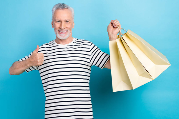 Photo portrait of funny old male hold shopping bags show thumb up dressed stylish striped garment isolated on blue color background. - Foto, afbeelding