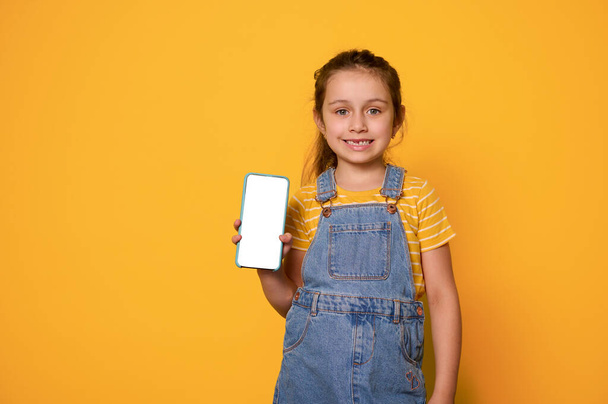 Adorabile ragazza vestita con t-shirt gialla e tuta in denim blu, mostrando un telefono cellulare intelligente con schermo bianco vuoto, sorridente con un sorriso allegro e dentato, isolato sfondo giallo studio - Foto, immagini