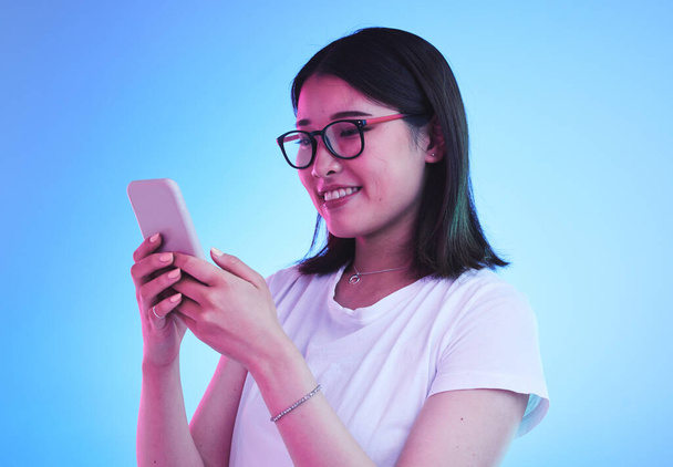 Phone call, reading an Asian woman in conversation on mobile app to contact person online in studio blue background. Connection, cellphone and young female chatting with smile for communication. - Photo, Image