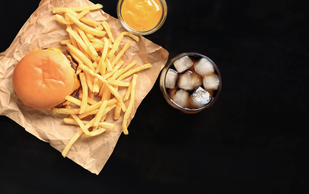 Hambúrguer de queijo e batatas fritas em papel marrom e mesa de madeira, vista superior. Hambúrguer, batatas fritas, cola com gelo num copo e mostarda. Fast food - Foto, Imagem