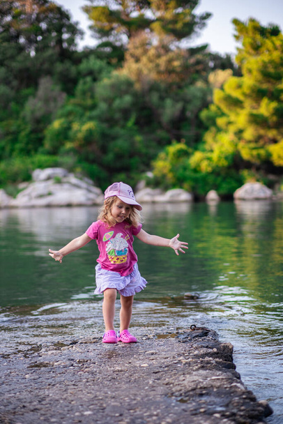 Dziewczynka w różowych ubraniach bawiąca się na kamiennym brzegu morza. Na tle są zielone drzewa i spokojna woda zatoki morskiej. - Zdjęcie, obraz