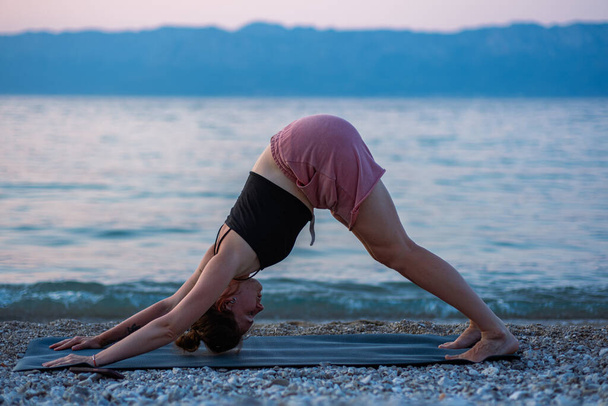 Une jeune fille en débardeur noir et short rose faisant du yoga sur la plage de la mer. Yoga au coucher du soleil surplombant la belle mer Adriatique et les rives vertes. Photo favorise un mode de vie sain, le sport, le yoga. - Photo, image
