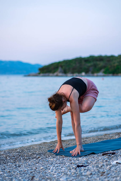 Ein junges Mädchen in schwarzem Tank-Top und pinkfarbenen Shorts beim Yoga am Meeresstrand. Yoga bei Sonnenuntergang mit Blick auf die wunderschöne Adria und die grüne Küste. Foto fördert einen gesunden Lebensstil, Sport, Yoga. - Foto, Bild