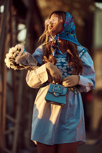 Urok kobiety ozdobionej wykwintną niebieską tradycyjną sukienką, niosącej niebieską torebkę i bukiet kwiatów, z wdziękiem spacerując po mieście o zachodzie słońca, tworząc hipnotyzującą scenę - Zdjęcie, obraz