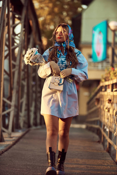 Urok kobiety ozdobionej wykwintną niebieską tradycyjną sukienką, niosącej niebieską torebkę i bukiet kwiatów, z wdziękiem spacerując po mieście o zachodzie słońca, tworząc hipnotyzującą scenę - Zdjęcie, obraz