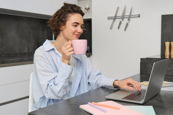 Beautiful girl working from home, drinking coffee and looking at laptop. Young businesswoman sits in kitchen and manages her business via computer. - Photo, Image