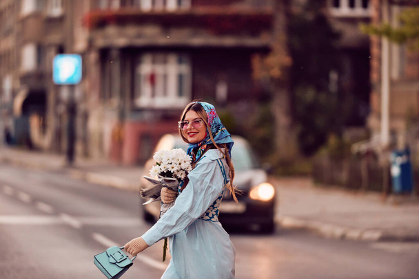 Encanto de una mujer adornada con un exquisito vestido tradicional azul, llevando un bolso azul y un ramo de flores, paseando con gracia por la ciudad al atardecer, creando una escena fascinante de - Foto, imagen