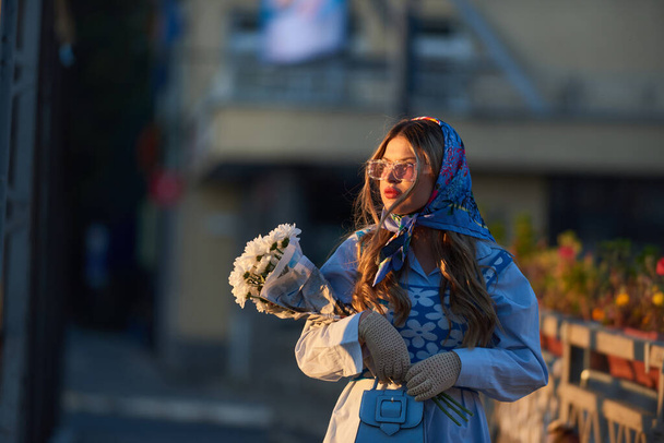 美しいブルーの伝統的なドレスに身を包んだ女性の魅力は、青いハンドバッグと花の花束を持ち、日没時に街を優雅に歩き、魅惑的なシーンを作り出します - 写真・画像