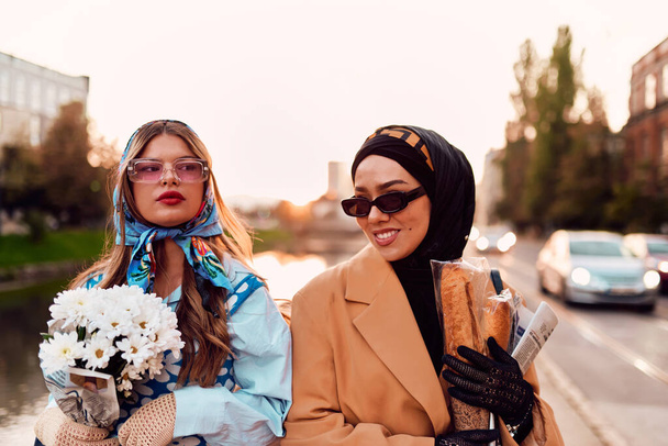 Pareja mujer una vistiendo un hijab y un vestido moderno pero tradicional, y la otra en un vestido azul y bufanda, caminando juntos por la ciudad al atardecer. Uno lleva un ramo y pan, mientras que el - Foto, imagen