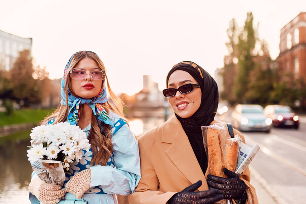 Een paar vrouwen met een hijab en een moderne maar traditionele jurk, en de andere in een blauwe jurk en sjaal, samen door de stad lopend bij zonsondergang. Men draagt een boeket en brood, terwijl de - Foto, afbeelding