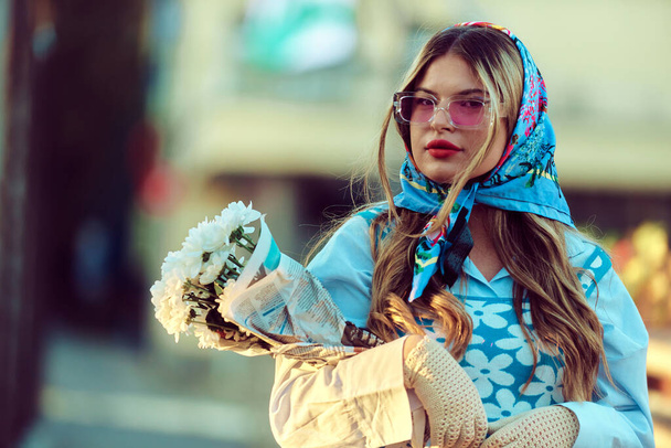 Encanto de una mujer adornada con un exquisito vestido tradicional azul, llevando un bolso azul y un ramo de flores, paseando con gracia por la ciudad al atardecer, creando una escena fascinante de - Foto, imagen