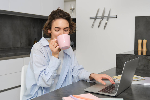 Εργαζόμενη γυναίκα που χρησιμοποιεί λάπτοπ, πίνει καφέ. Κορίτσι με φλιτζάνι τσάι, μελέτη από hyome, κάθεται κοντά στον υπολογιστή στην κουζίνα, προετοιμασία για τις εξετάσεις. - Φωτογραφία, εικόνα