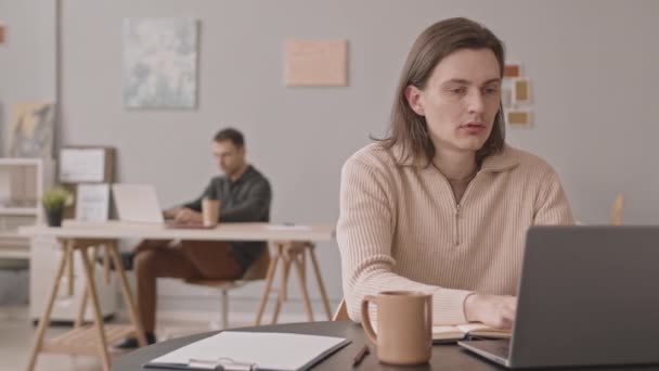 Joven hombre caucásico escribiendo en el ordenador portátil mientras trabaja en la oficina con un colega masculino - Imágenes, Vídeo