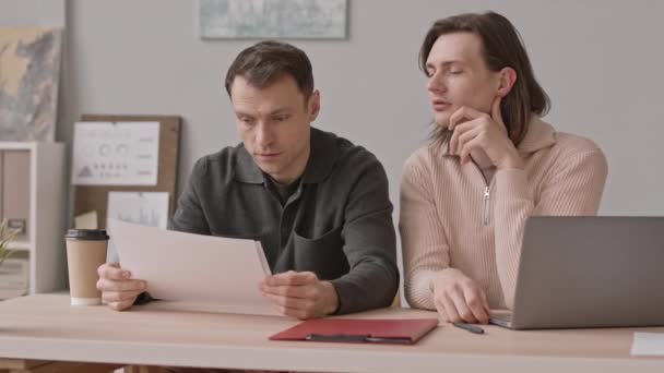 Καυκάσιος ομοφυλόφιλος ζευγάρι κοιτάζοντας μέσα από τα έγγραφα των επιχειρήσεων, ενώ κάθεται στο γραφείο μαζί εργάζονται στη σύγχρονη εταιρεία μαζί - Πλάνα, βίντεο