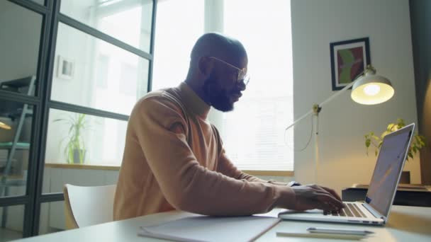 Μεσαία λήψη του Αφροαμερικανού επιχειρηματία πληκτρολογώντας σε φορητό υπολογιστή και κρατώντας σημειώσεις κατά τη διάρκεια της εργασίας στο γραφείο - Πλάνα, βίντεο